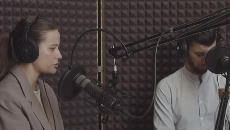 una joven elegante con el pelo recogido y vestido con traje habla sobre un tema interesante en un estudio de radio mientras un joven escucha a su lado