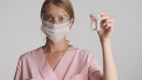 caucasian female doctor in safety goggles and mask holding flask with pills.