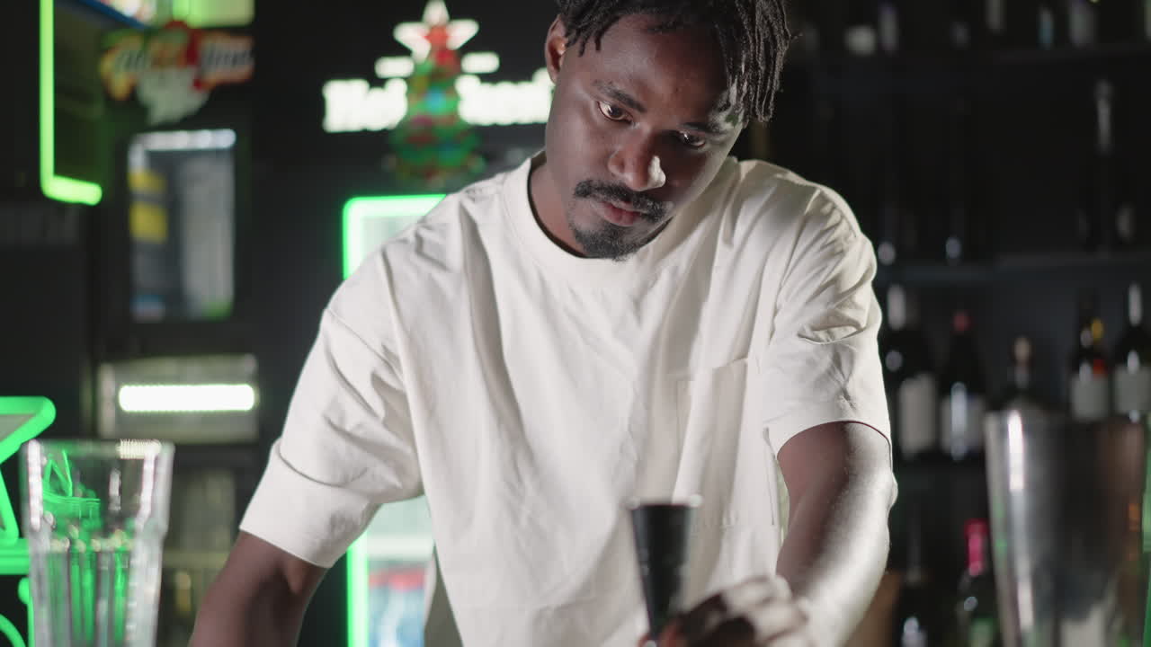 Bartender pouring whiskey from bottle into cocktail shaker with focused expression, preparing drink at modern bar with neon lighting in vibrant atmosphere