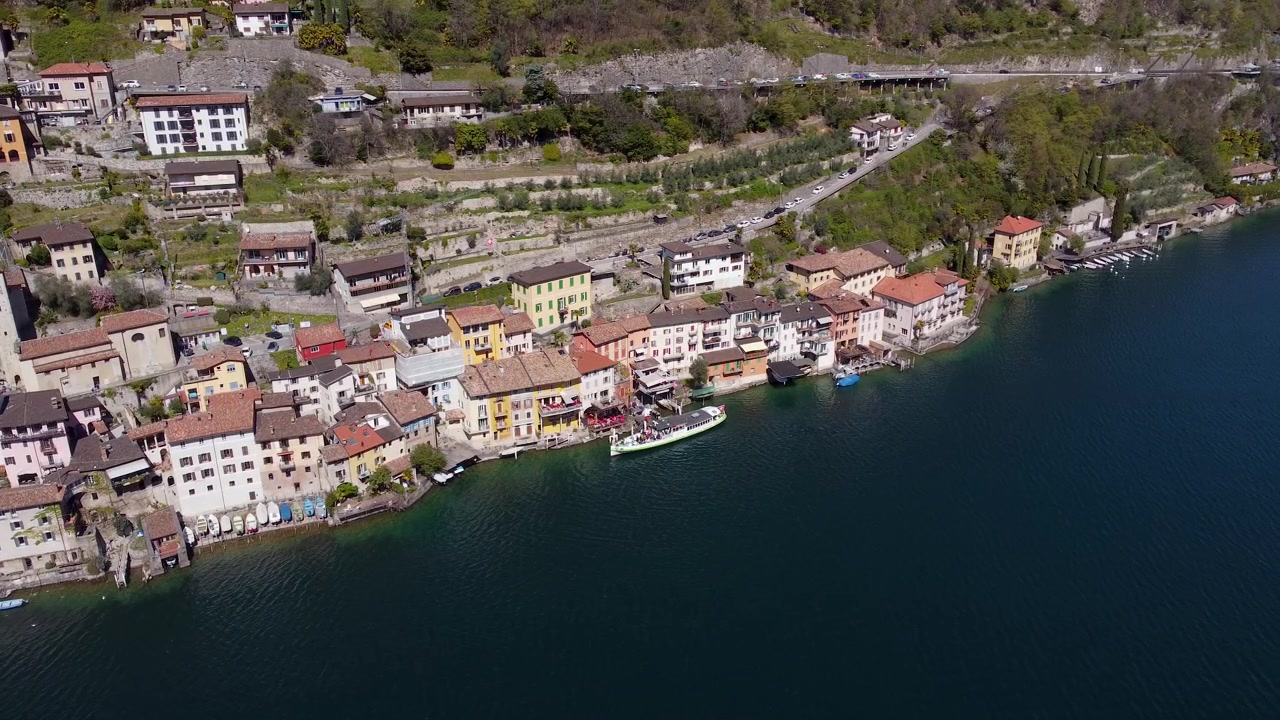 Case color pastello fiancheggiano il villaggio lacustre di Gandria, sul Lago di Lugano, Svizzera. Un traghetto bianco attracca vicino a vigneti terrazzati sul fianco della collina, sopra una strada costiera tortuosa. Ampia ripresa aerea con drone.