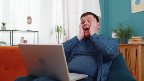 man sitting on a couch surprised by something on his laptop.