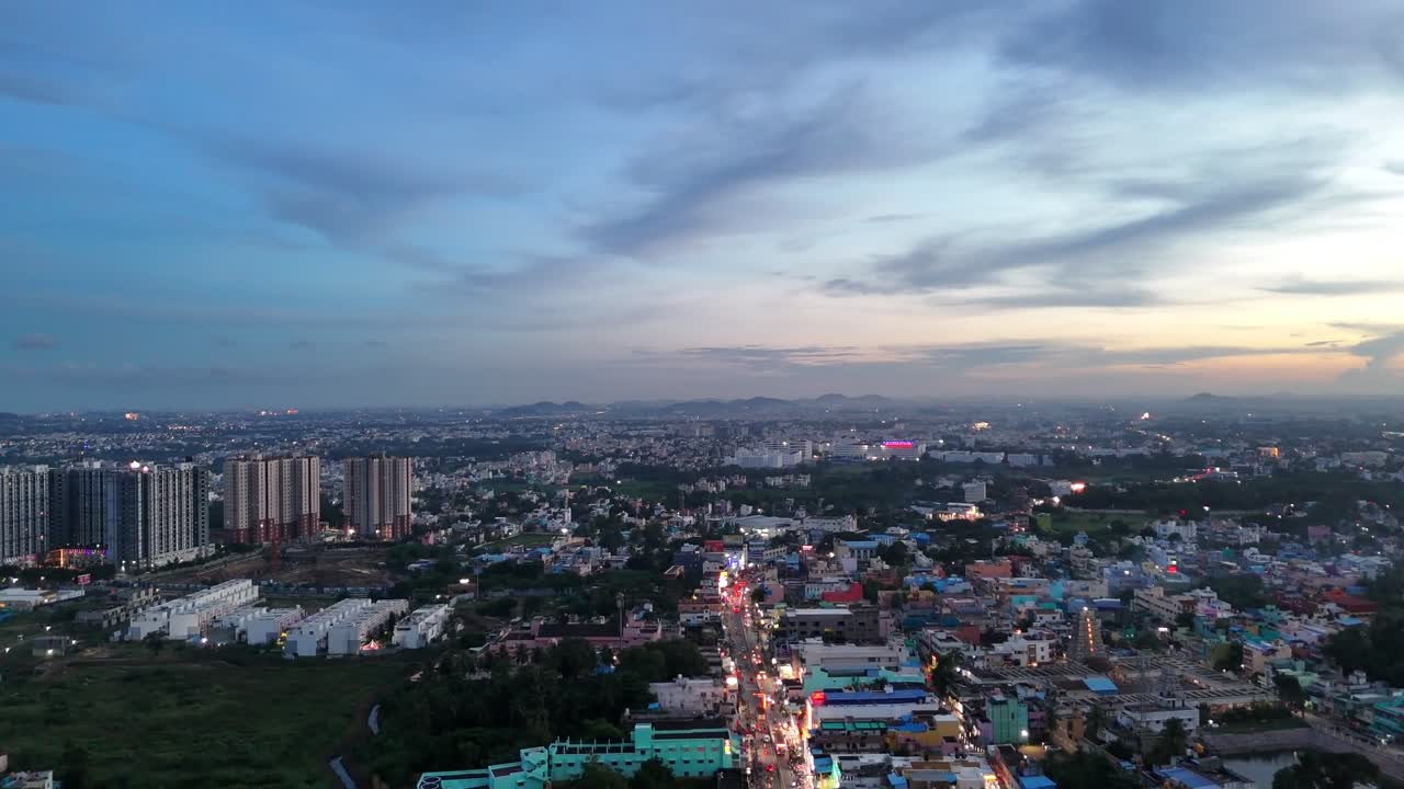 Il paesaggio urbano di Chennai all'alba mostra strade antiche e fitte in primo piano e alti palazzi residenziali. I lampioni stradali e le scie del traffico illuminano la scena, con colline distanti visibili all'orizzonte.