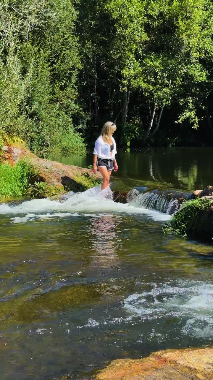 A blonde caucasian woman crossing a fast flowing river between the rocks at a small waterfall