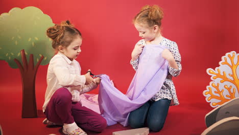 two little girls are playing with a lilac cloth on a pink studio background. one of girl is cutting fabric by scissors in her hand