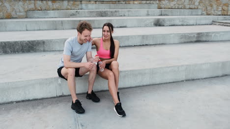 glückliches sportliches paar, das draußen auf treppen sitzt, wasserflaschen hält und miteinander redet, während es während seiner laufeinheit in der stadt eine pause macht