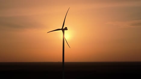 Molino-De-Viento-De-Toma-Aérea-Erguido-Contra-El-Fascinante-Telón-De-Fondo-De-Una-Ardiente-Puesta-De-Sol-Naranja,-Primer-Plano-Estilo-Country-Jour,-Proyectando-Un-Contorno-Dramático-De-Las-Aspas-Del-Molino-De-Viento