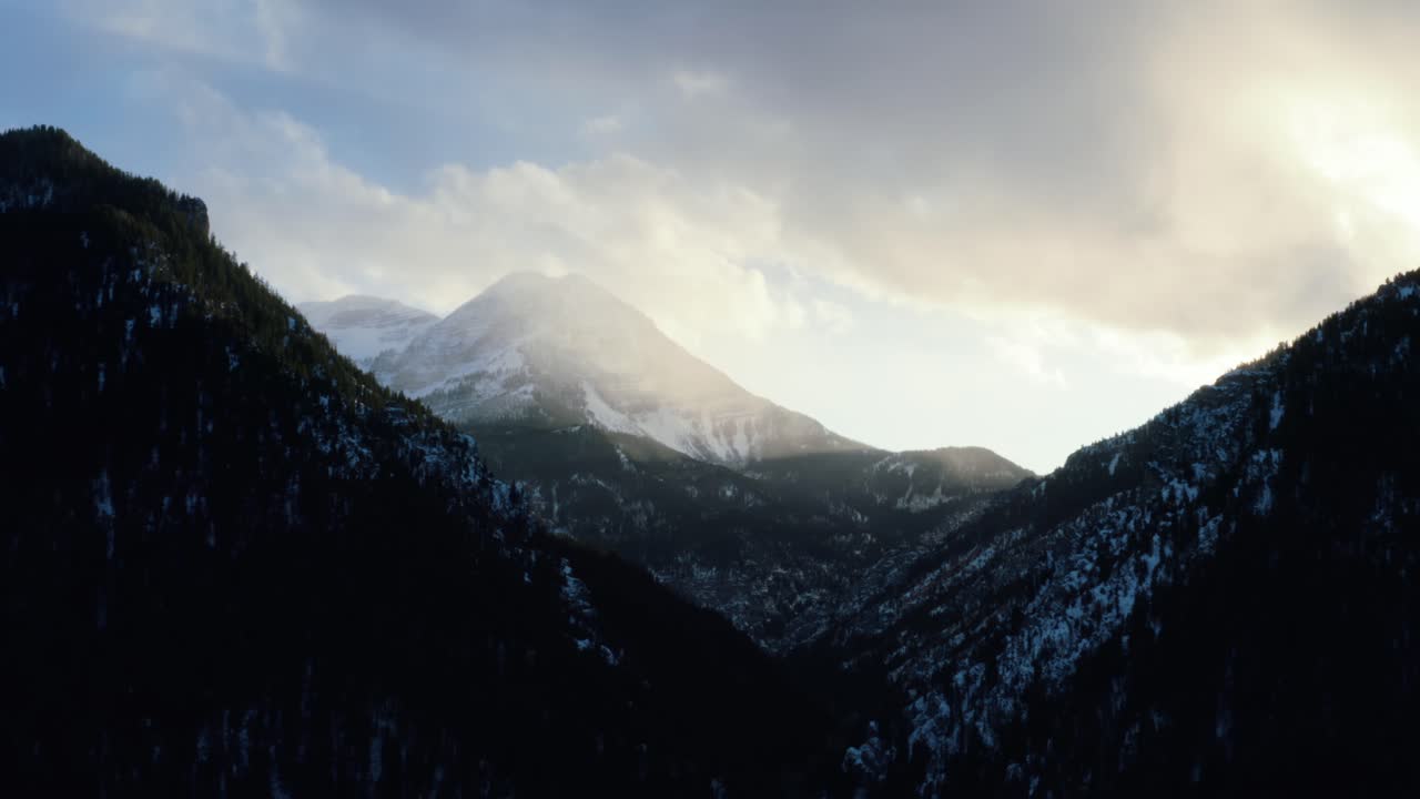 Static aerial drone shot of a winter landscape of Mount Timpanogos in the background surrounded by a pine tree forest during sunset from the frozen Tibble Fork Reservoir in American Fork Canyon, Utah