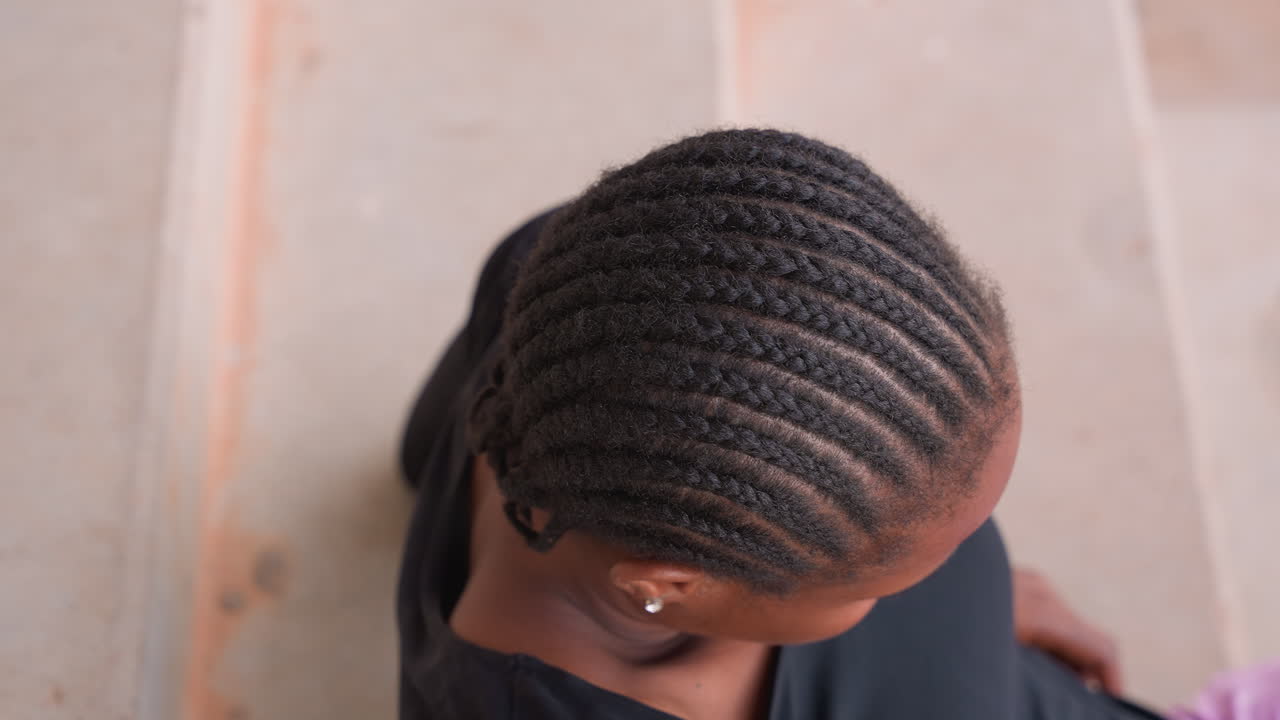 Femme noire avec des tresses collées assise sur des escaliers en béton, portant un haut noir et un pantalon violet, pose détendue et regard vers le haut, lumière douce du jour et marches texturées, portrait aérien rapproché capturant le calme.