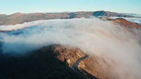 mountain road winding through a sea of clouds, early morning light touches peaks, aerial shot