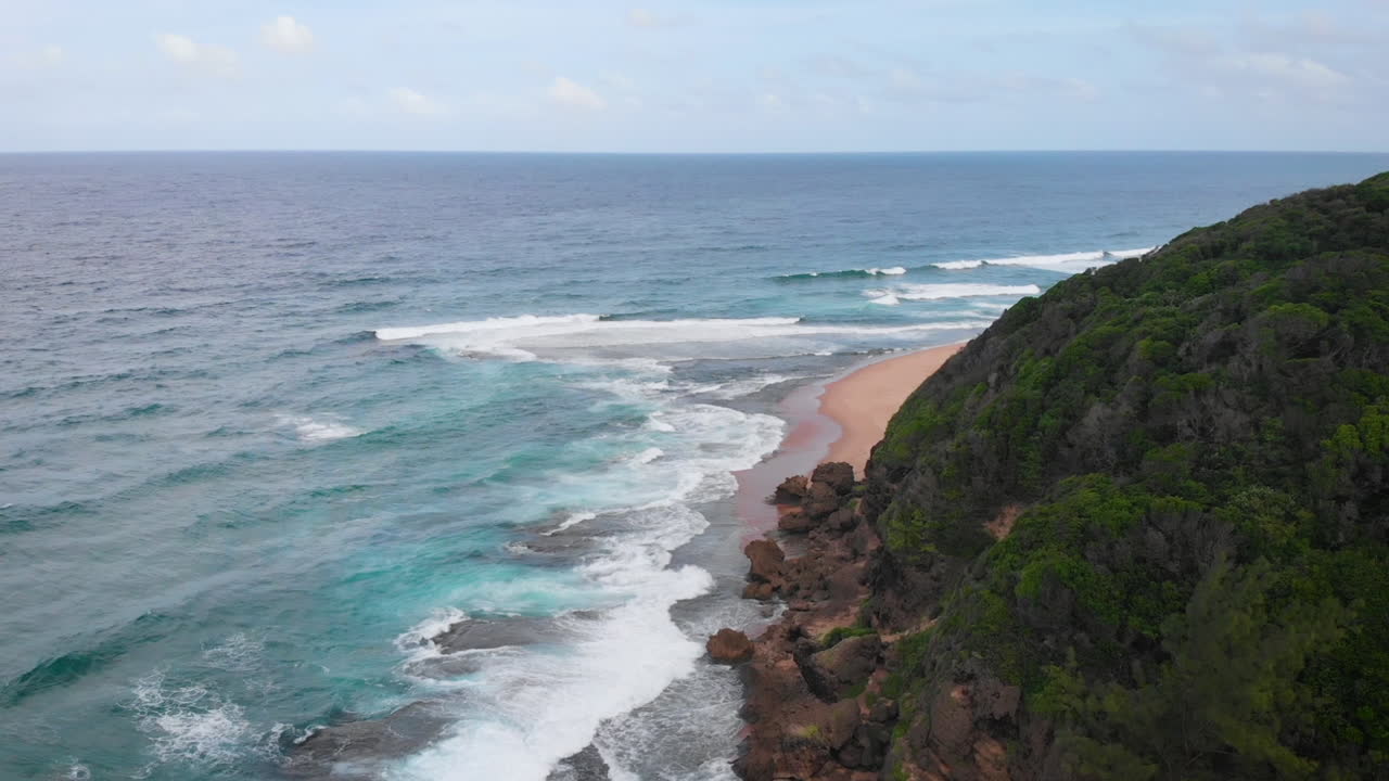 Vista aerea dell'oceano che incontra la parete rocciosa su un'isola tropicale alberata con la marea in arrivo.