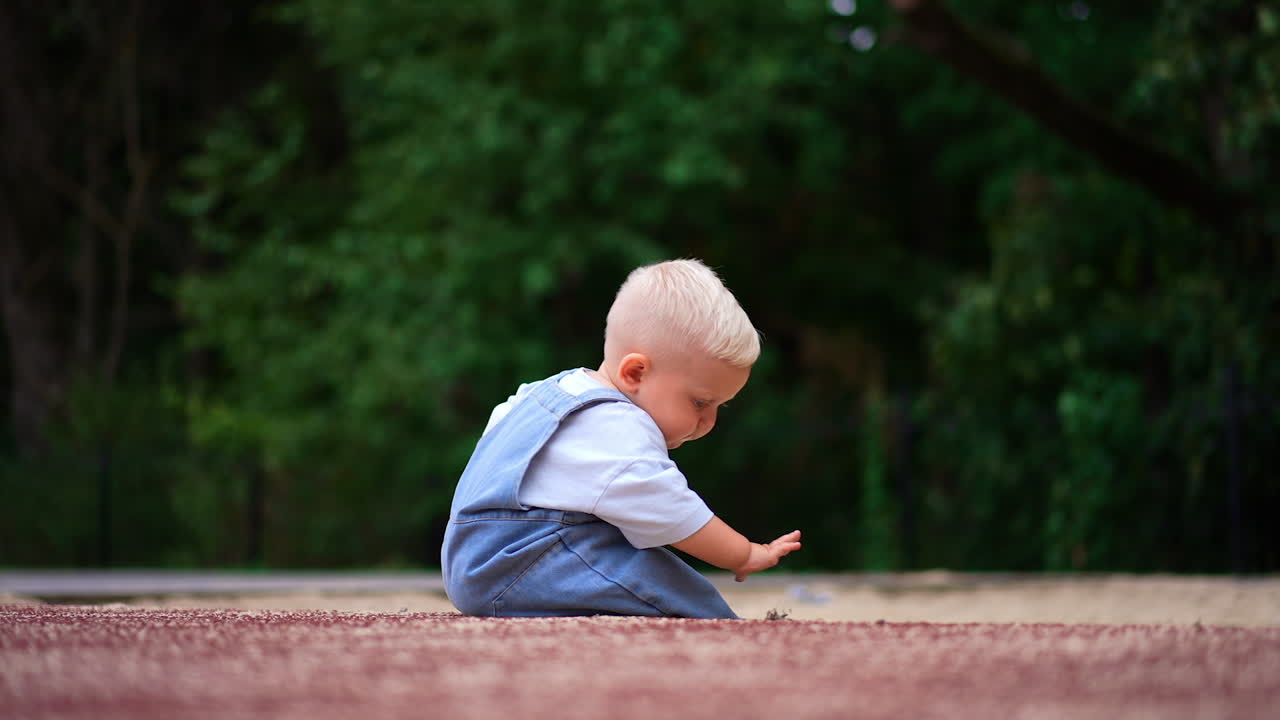 Small kid sitting on the ground outdoors. Curious kid plays with sand in summer. Blurred nature backdrop.