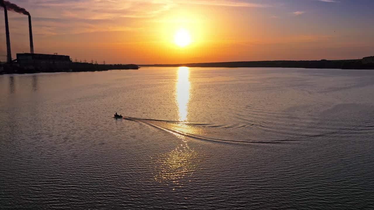 Aerial view over the water and motor boat floating at sunset. Beautiful view on the setting orange sun that reflects in water. Boat on the river moves to the industrial factory.