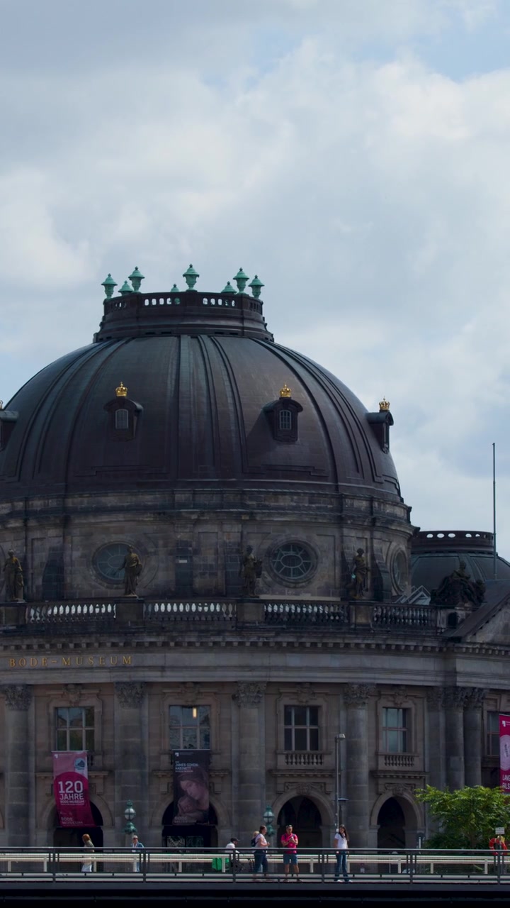 La cámara se desplaza por la cúpula del Museo Bode, capturando a peatones y un cielo de verano nublado en Berlín.