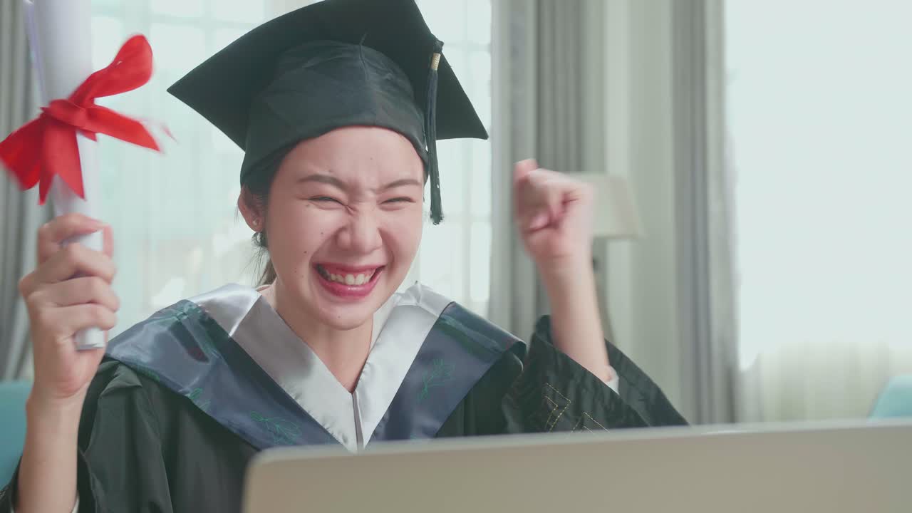 Asian Woman Celebrating With University Certificate To The Family During An Online Video Call. Pretty Female Graduate Wearing A Graduation Gown And Cap Sitting On The Living Room