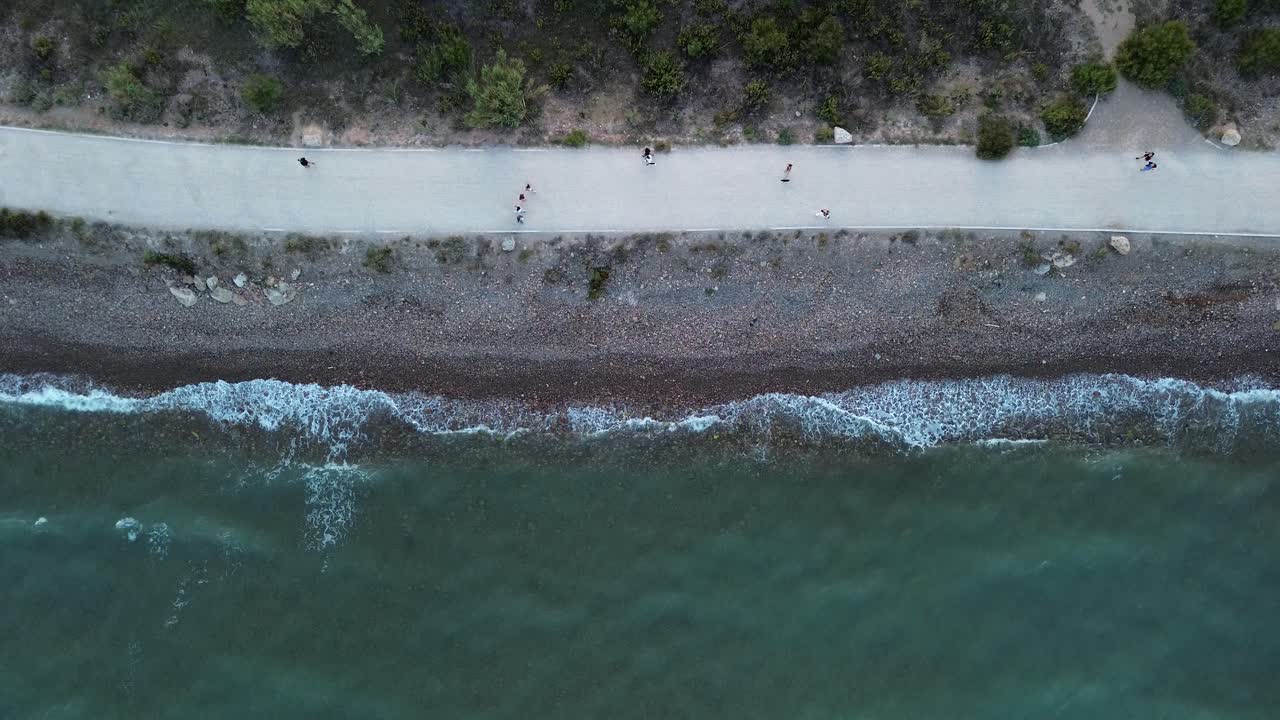 vista de arriba hacia abajo de un dron de una playa serena con olas golpeando suavemente la costa al atardecer, mientras la gente camina y hace ejercicio a lo largo de la costa