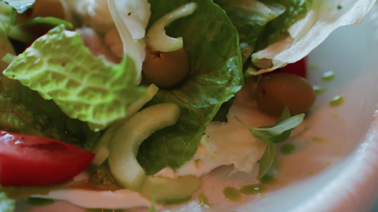Primo piano di insalata romana, olive e pomodori, texture vivaci con condimento che cola, riccioli di cipolla affettata, nido di pomodorini ciliegino, prospettiva di ciotola tenuta in mano, allestimento cucina casual, luce naturale brillante