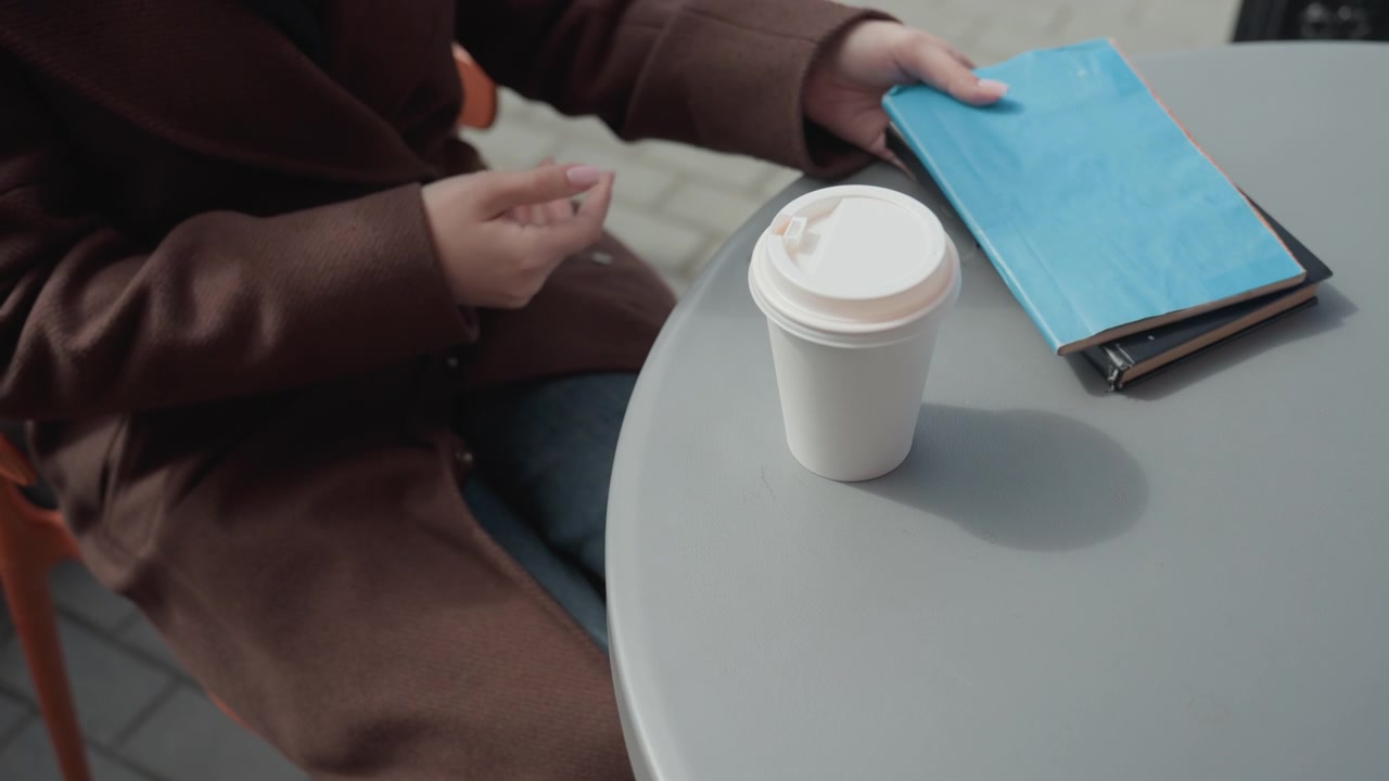 Una donna si avvicina a un ambiente esterno con tavoli e sedie. La donna estrae una sedia, si siede al tavolo e poi vi appoggia sopra una tazza di caffè prima di prendere il telefono