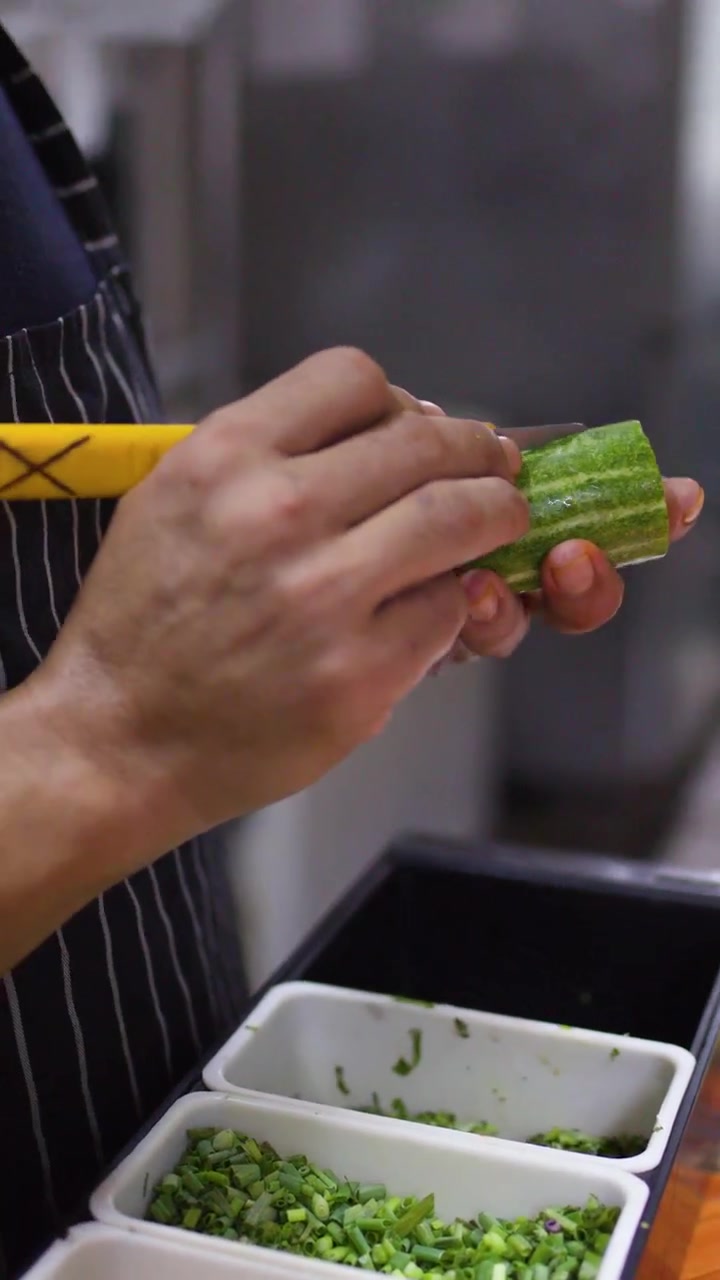 Primo piano delle mani di uno chef che affettano abilmente un cetriolo con un coltello in una cucina commerciale.