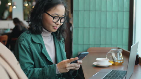 Asiatische-Frau,-Die-Im-Café-Nachrichten-Am-Telefon-Sendet