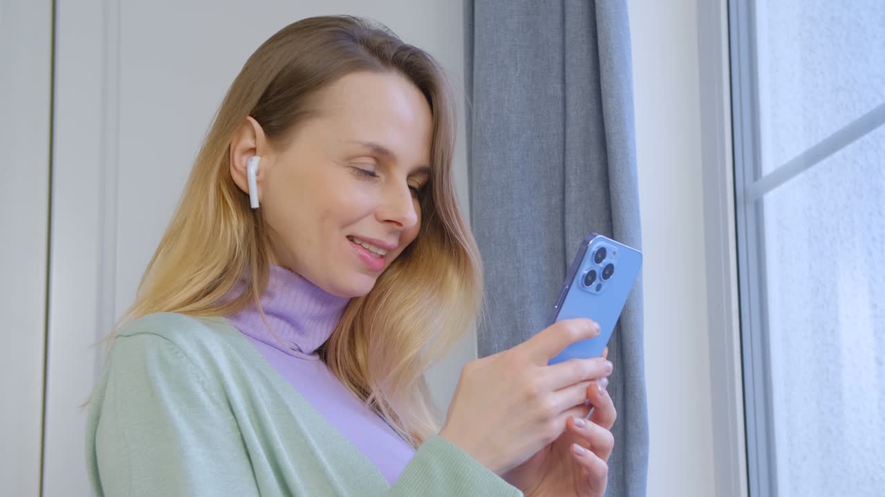 Donna bianca felice che ascolta musica in cuffie wireless e digita un messaggio sul cellulare. Allegra donna caucasica sui 40 anni che usa un moderno smartphone blu per la comunicazione online e l'intrattenimento.