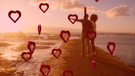 animation von roten herzen über ein verliebtes paar, das sich am meer an der hand hält.
