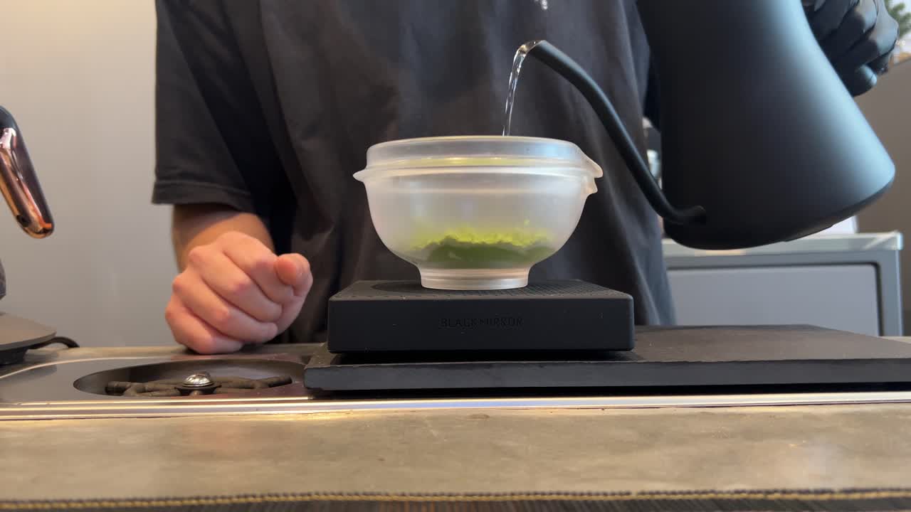 A barista pours hot water into a bowl of matcha powder, creating tea in a Phuket cafe setting