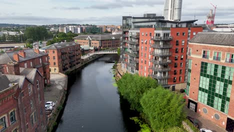 drone camera glides above river aire, revealing victorian red-brick buildings, modern apartments, and lush riverside greenery in central leeds under daylight