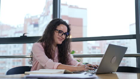 Beautiful woman working at computer and smiling. Female worker in a good mood works in an office. Workplace in big light office with large windows.