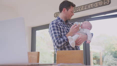 Working-father-using-laptop-at-home-whilst-feeding-baby-son-sitting-on-his-knee--from-bottle---shot-in-slow-motion