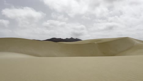 majestic sand dunes stretch across the viana desert on boa vista, cape verde, beneath a dramatic, cloud-laden sky. a stunning blend of textures and vast open space, mountains in background