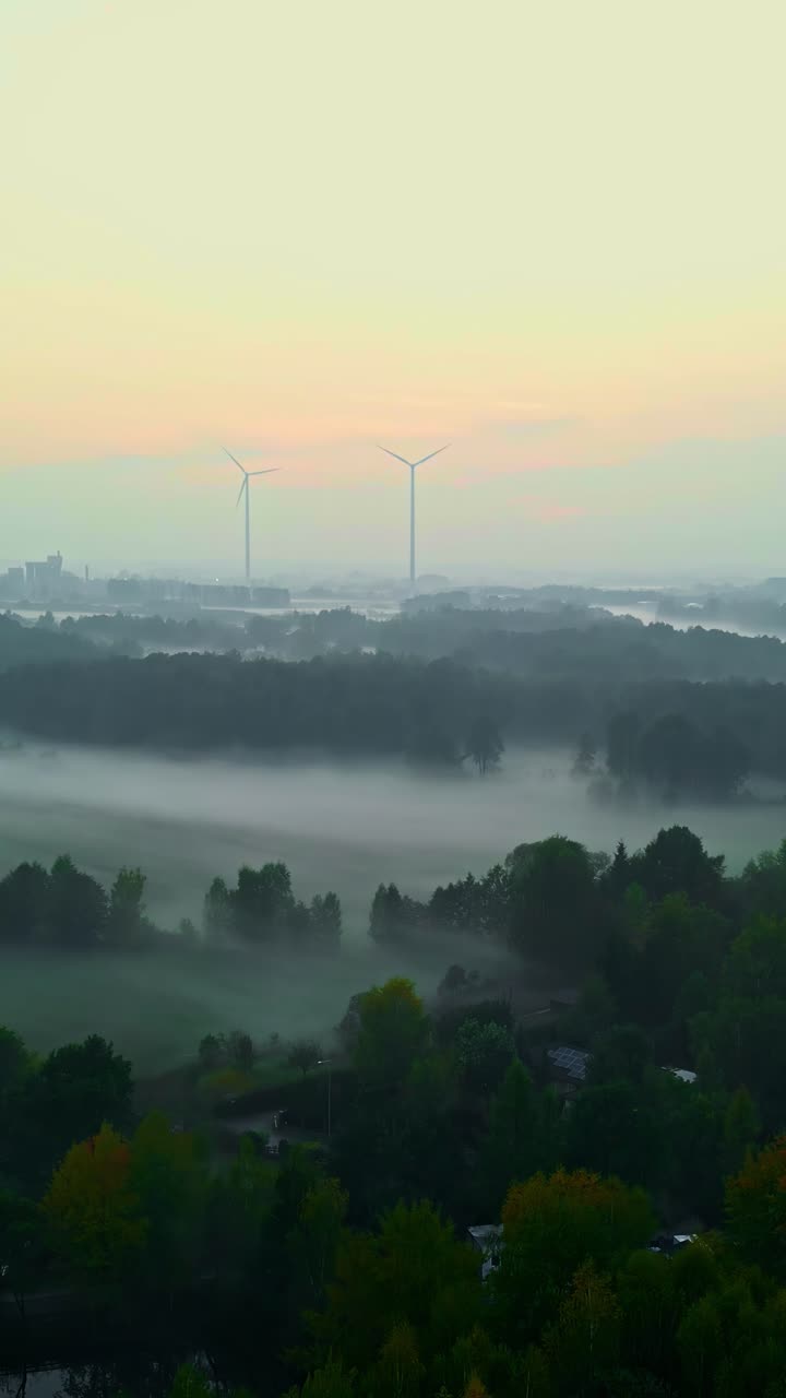 Vertical aerial view of a misty forest landscape in Latvia at dawn with two distant wind turbines and a city skyline emerging from the fog