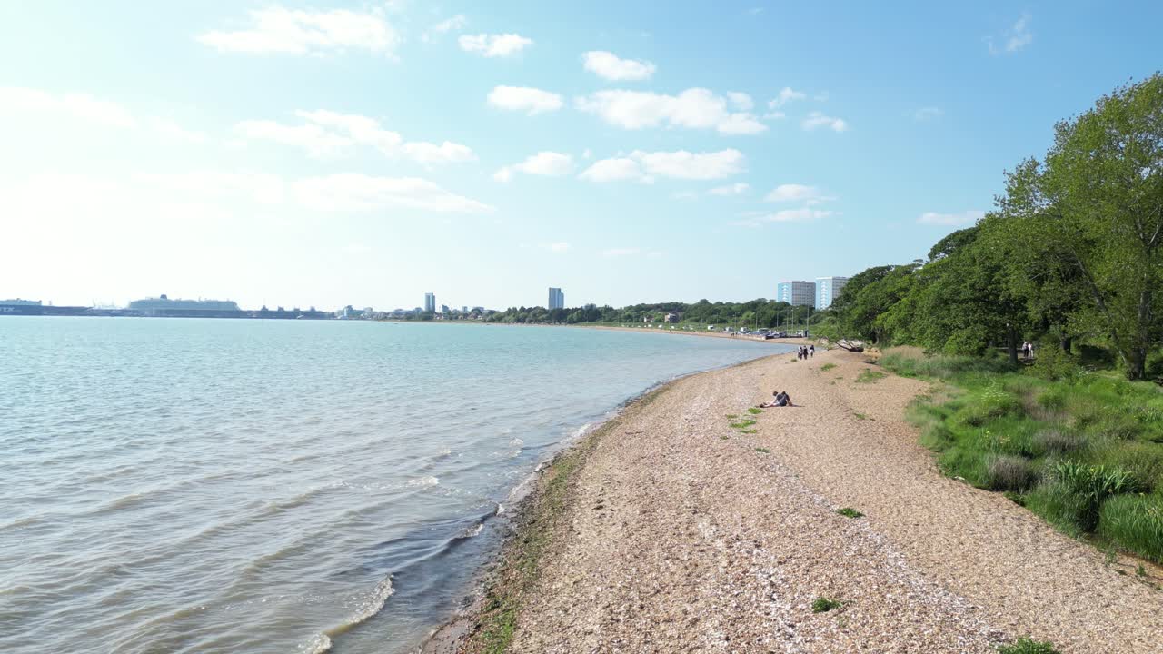 Wide aerial view of calm waterfront with pebbled beach, green trees, and distant city skyline under blue sky in Canada, showing peaceful coastal environment and quiet natural urban contrast