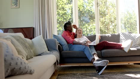 happy diverse couple relaxing on couch using laptop in living room, copy space, slow motion