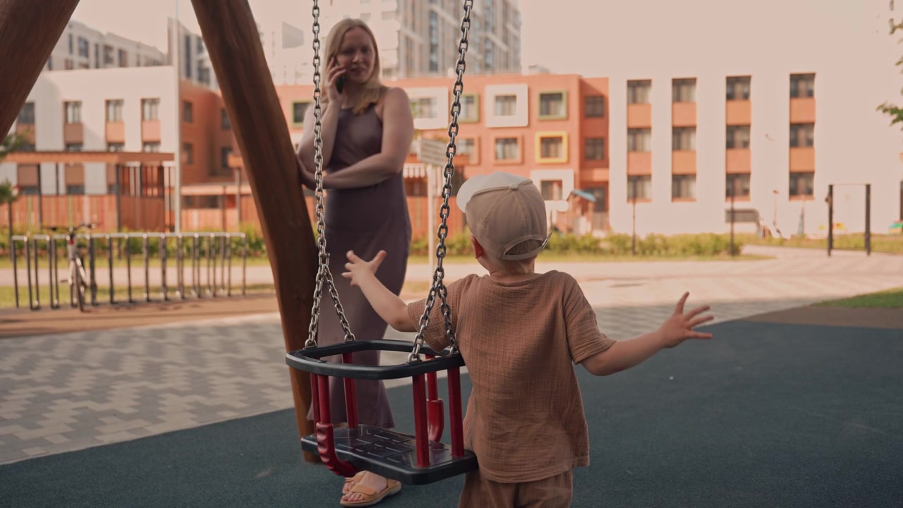 Un bambino esplora l'altalena in un parco giochi all'aperto mentre una donna sta nelle vicinanze parlando al telefono. L'ambientazione presenta moderni edifici residenziali in una luce diurna soffusa con superfici in cemento e gomma.