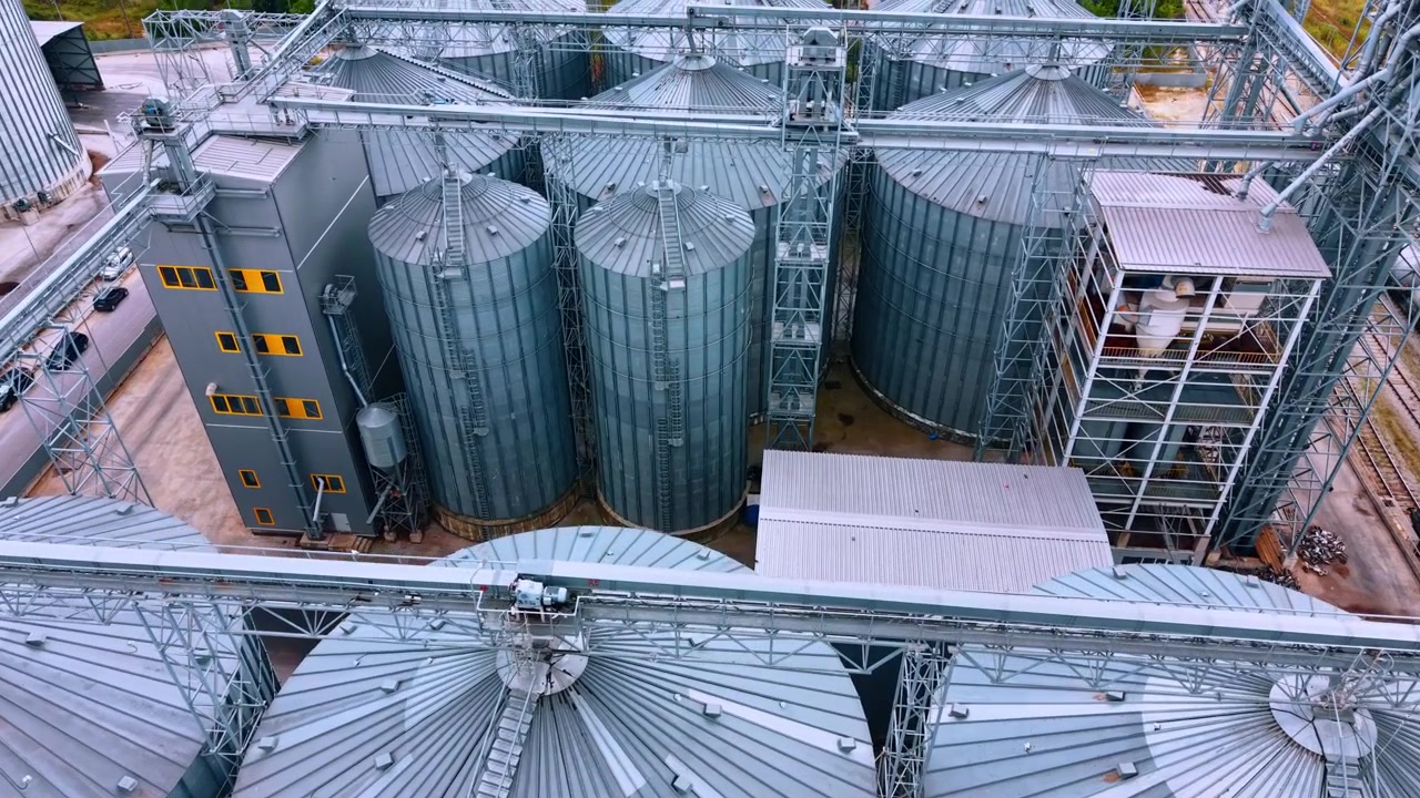 Conveyor network above industrial grain silos. Elevated view of corrugated metal bins, access ladders, processing towers and tanker wagons beside rail tracks.