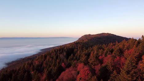 Tiro-De-Dron-De-Una-Montaña-Cubierta-En-Un-Bosque-De-Otoño-Con-Vistas-A-Un-Mar-De-Nubes-Al-Atardecer