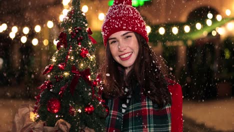 vista de cerca de una mujer caucásica con abrigo rojo sosteniendo un árbol de navidad y sonriendo a la cámara en la calle mientras nieva en navidad