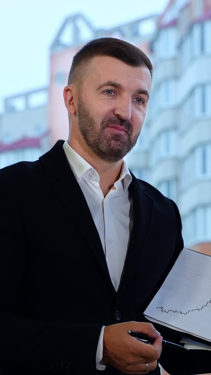 Talkative man shows a graph and comments it to his colleagues. Positive man in official suit explaining charts at office. Big window with house at the backdrop. Vertical video