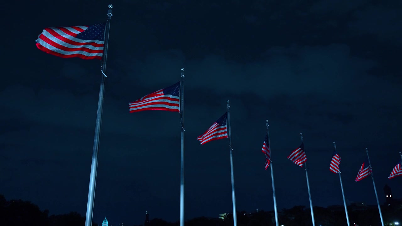 American flags circling the Washington Monument at night, Washington DC. Wide angle of illuminated Capitol dome visible on the distant horizon.
