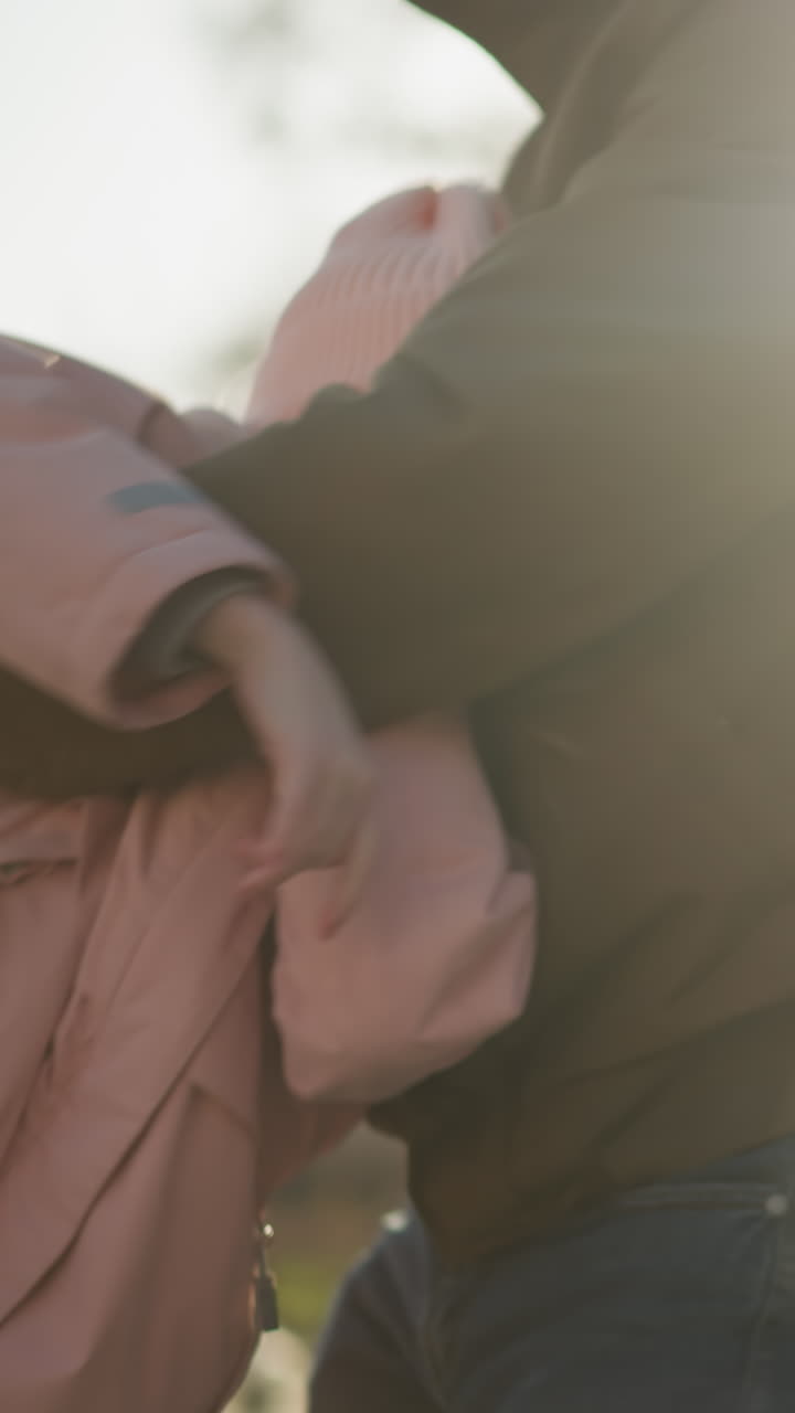 una niña con una gorra rosa y una chaqueta siendo girada juguetón por un hombre con una chaqueta marrón y pantalones vaqueros azules. la escena captura un momento alegre con la cálida luz del sol filtrándose a través