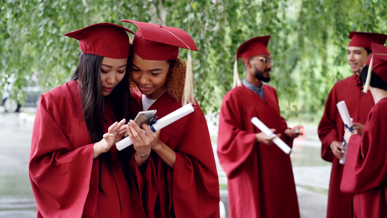 Attraktive unge kvinder, glade dimittender, ser billeder på smartphone og taler på dimissionsdagen, mens deres medstuderende snakker i baggrunden.