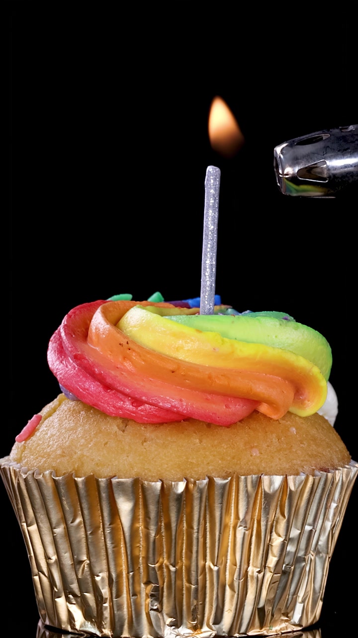 Una mano accende una candela di compleanno su un cupcake glassato arcobaleno su sfondo nero con illuminazione brillante e uniforme.