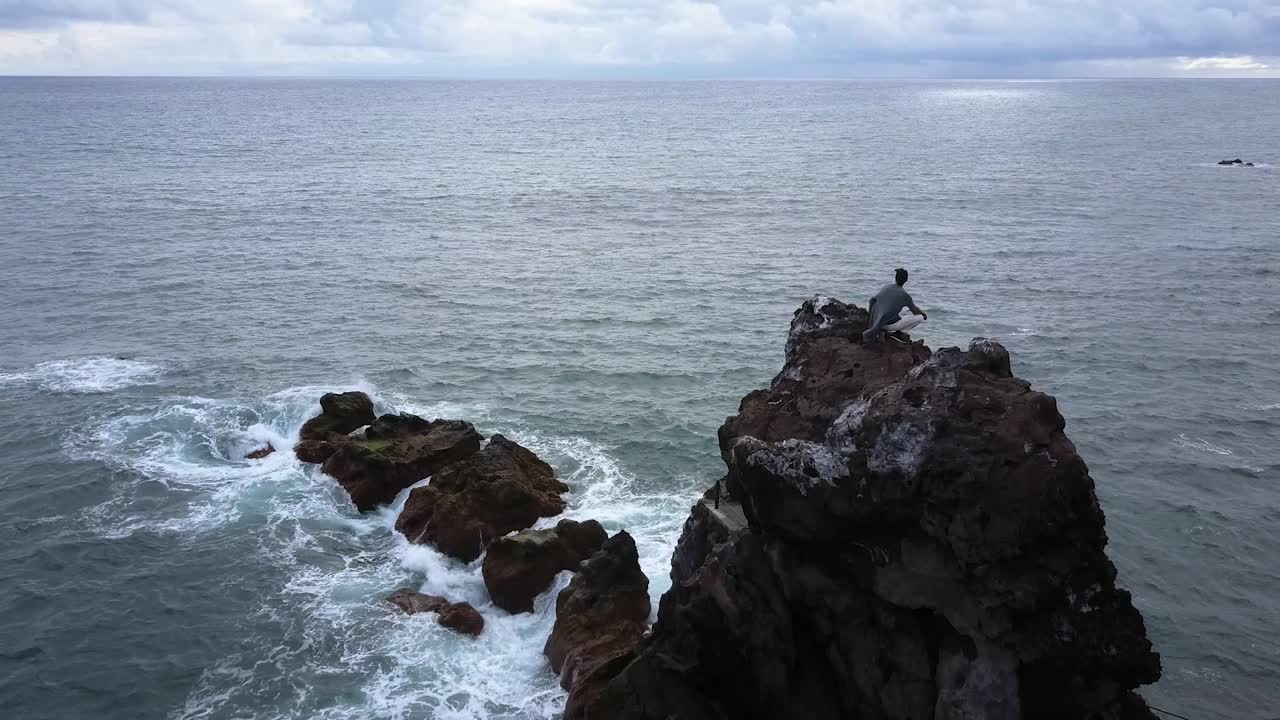 isla de madeira, portugal - hombre sentado en la cima de un acantilado mirando las olas del mar salpicando en las rocas - tiro aéreo de drones