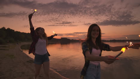 cheerful male and female friends are running along the beach at sunset holding sparkling fireworks and runaway lights in slow motion. dancing and sunset party on the beach