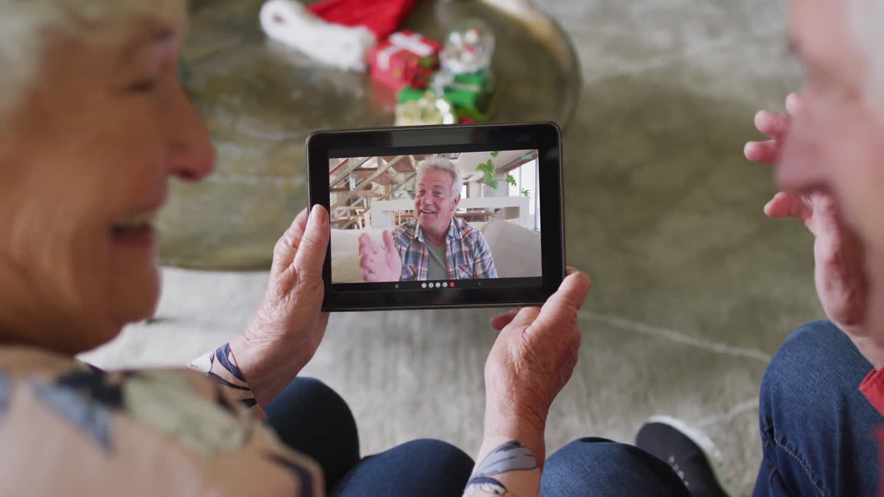 Coppia anziana caucasica sorridente che usa un tablet per una videochiamata di Natale con un uomo sullo schermo.