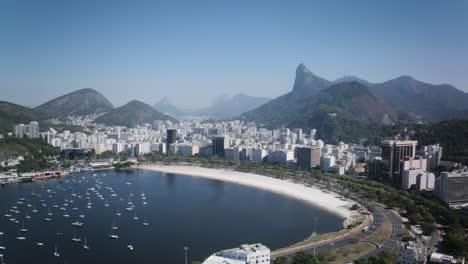 Ariel-Lapso-De-Tiempo-De-La-Playa-De-La-Bahía-De-Botafogo-Con-Veleros-Y-Tráfico-Circulando-Durante-El-Día-Con-La-Estatua-Del-Cristo-Redentor-Al-Fondo-En-Río-De-Janeiro