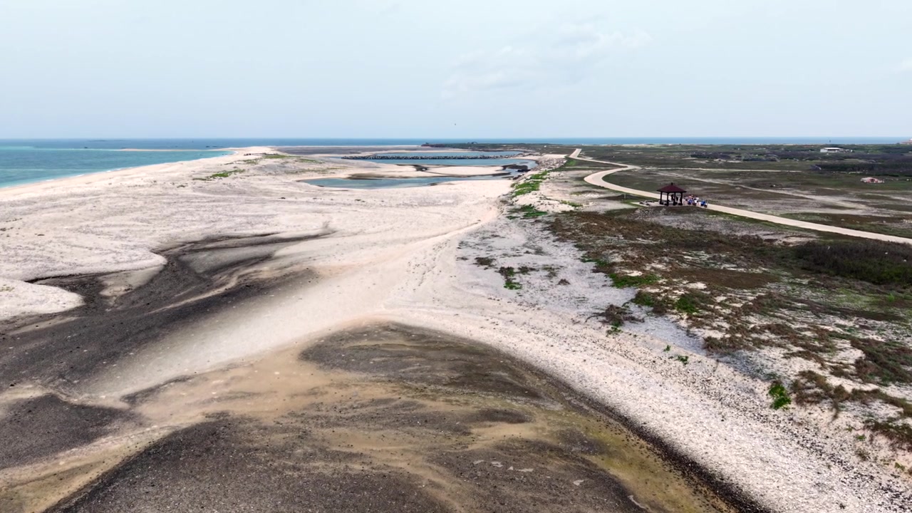 Mørke tidevandskanaler strækker sig hen over en hvid sandstrand på Jibei-øen i Penghu, Taiwan, med en lille rød pavillon og en kyststi, der strækker sig mod en fjern havnemole. Luftfoto fra drone.