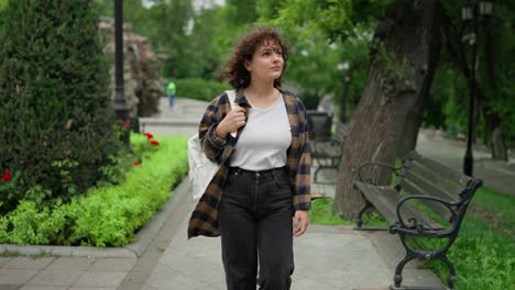 A-happy-brunette-girl-with-curly-hair-in-a-checkered-shirt-and-a-white-T-shirt-with-a-white-backpack-walks-through-the-park-after-her-classes-at-the-university