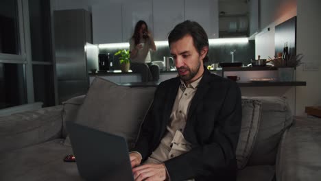 A-confident-brunette-man-with-stubble-in-a-black-jacket-and-beige-shorts-sits-on-the-sofa-and-works-at-a-gray-laptop-while-his-brunette-girlfriend-in-a-beige-T-shirt-sits-in-the-kitchen-and-drinks-tea