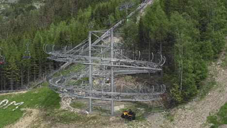 rotating aerial 4k shot of a big loop of an extreme outdoor roller coaster ride in the mountains of dolní morava, czech republic, with trees and a cableway in the background